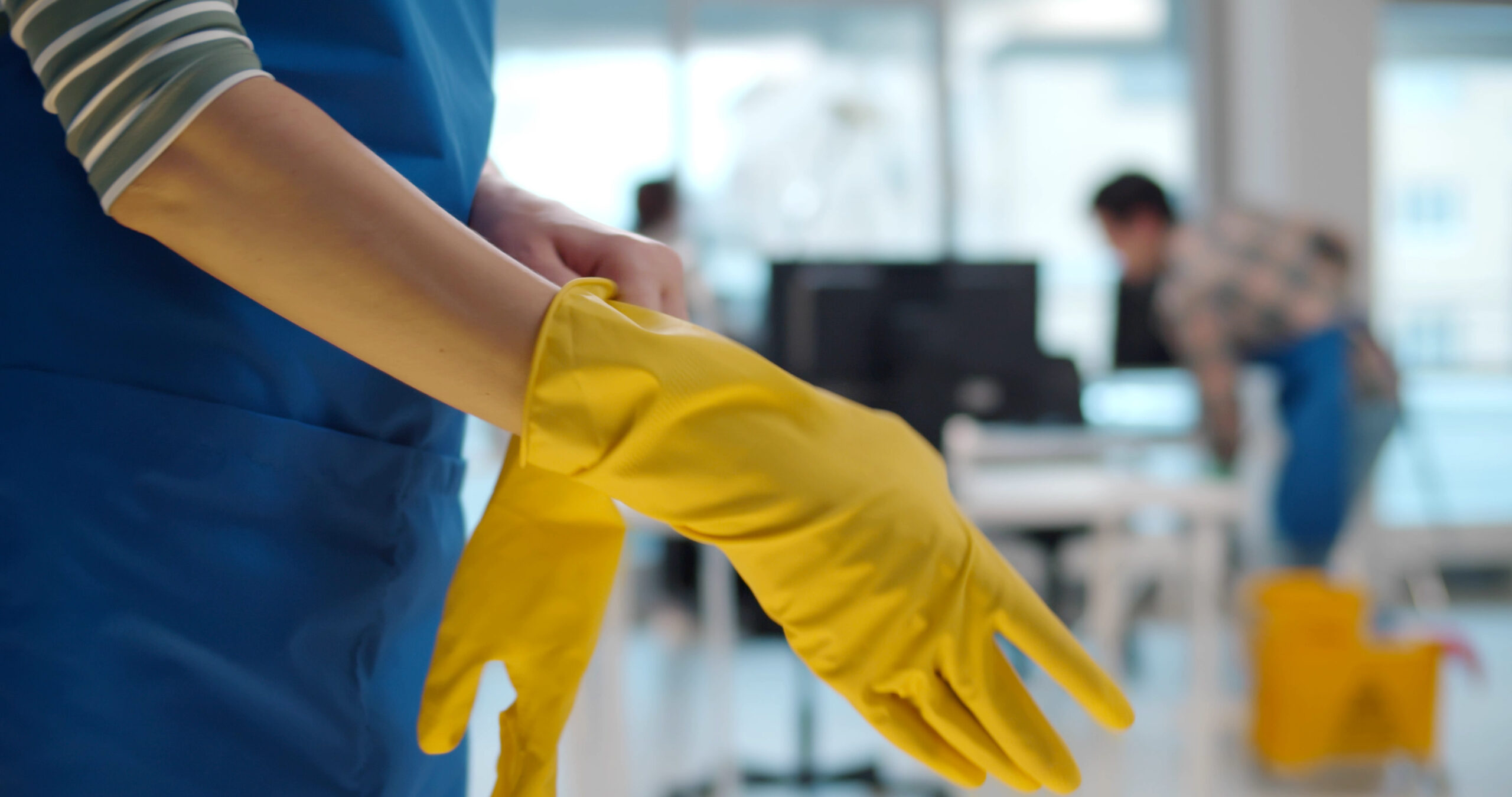 Close up of woman maid putting on rubber gloves with colleagues cleaning office on background